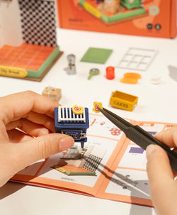 A person assembling Afternoon Baking Time Mini Diy House kit with tools. Simplified assembly, no painting needed. Dimensions: 5.3 x 2.2 x 5.9 in. Reflects Strangecat Toys' blind box and art toy store essence.