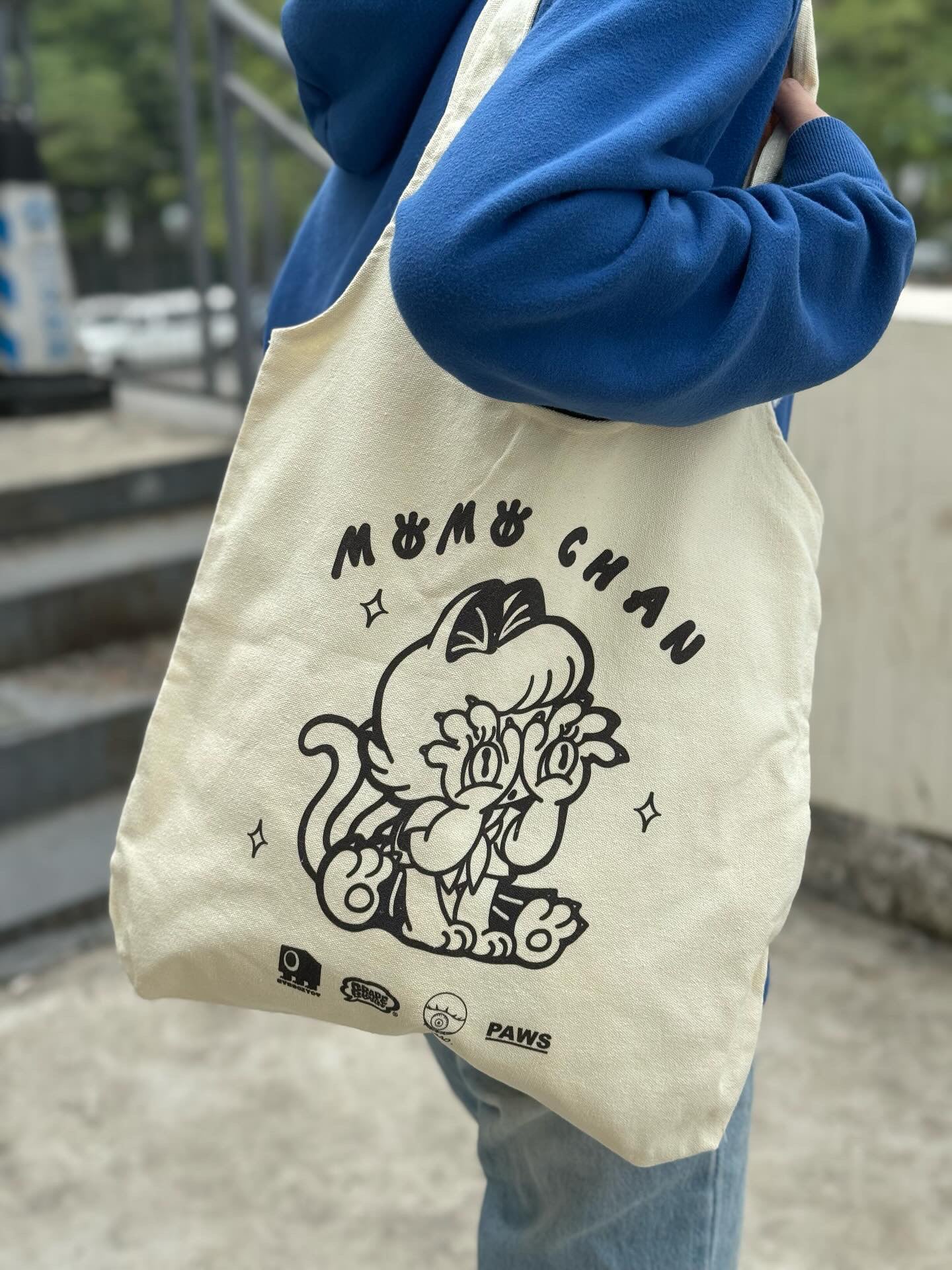 A person holding a Momochan Tote Bag by Abao x Grape Brain featuring a cartoon cat design. Preorder available, ships Aug 2024.