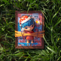 Toy figurine of child in Santa hat wrapped in tape on grass, with glass and symbol nearby.