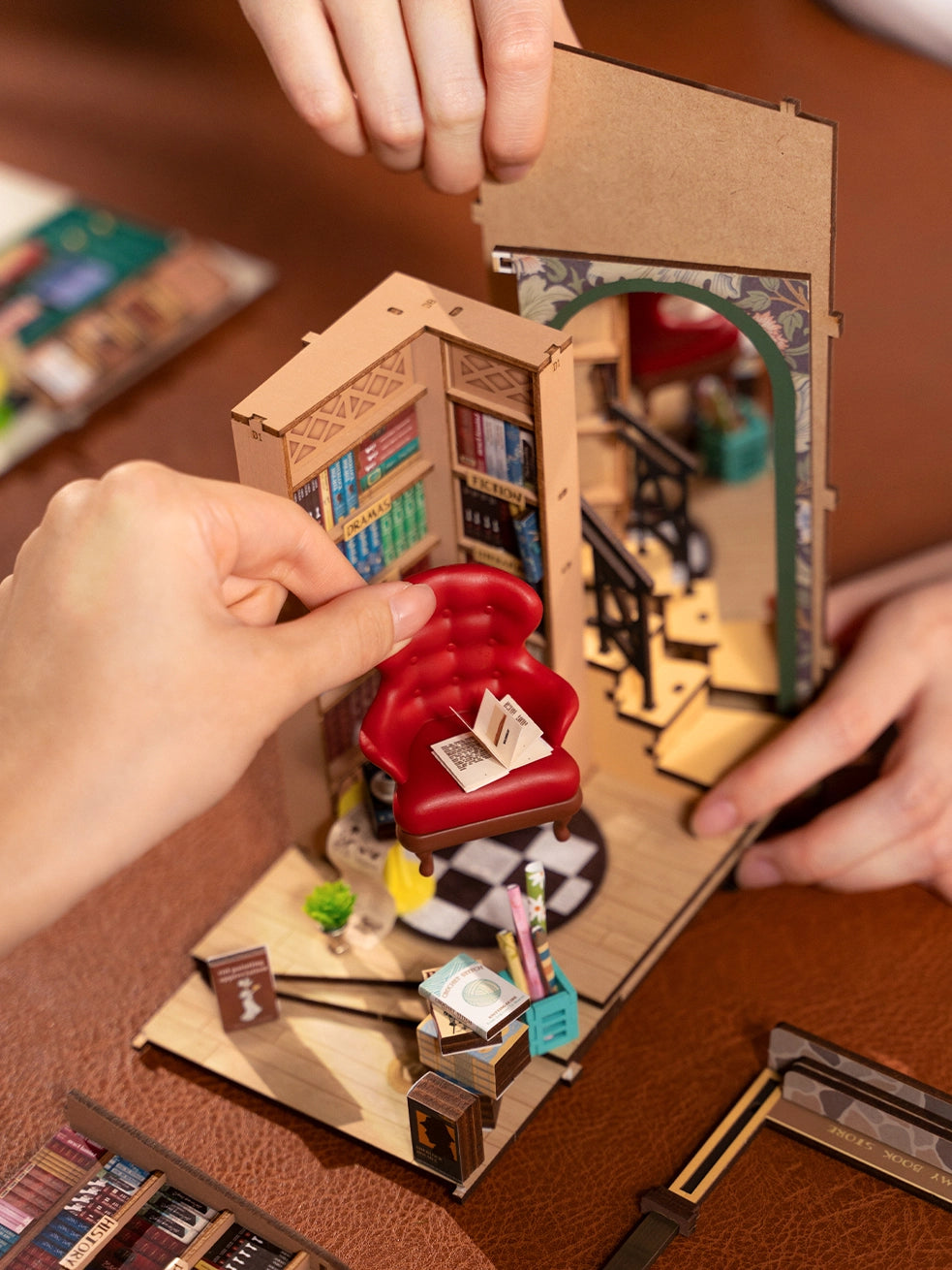 A hand holds a miniature bookcase with a book on a red chair, emphasizing creativity and assembly. Book Nook Kits For Adults - Bookstore from Strangecat Toys.