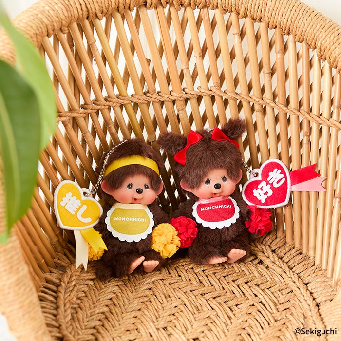 Happy Color Monchhichi Keychain nestled among stuffed animals in a wicker basket, featuring a heart tag and ball chain for easy portability.