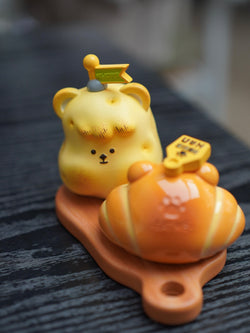 Yellow bear figurine with croissant hat on a wooden board, part of the Hanhan Bread series by Unknown Island.