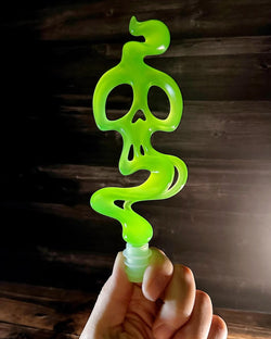 A hand holding a green resin bottle topper with a skull shape, silicone base for wine safety. Glow in the dark. 7 1/4” tall, 2 3/4” wide. Preorder - Ships Late Sept 2024.