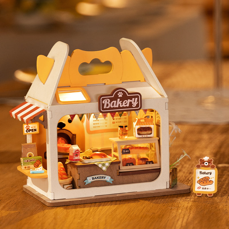 Toy bakery model, Teddy’s Breadbox Diy Miniature House, featuring illuminated display case, bread, brown sign with white text, and bear label, on a table.