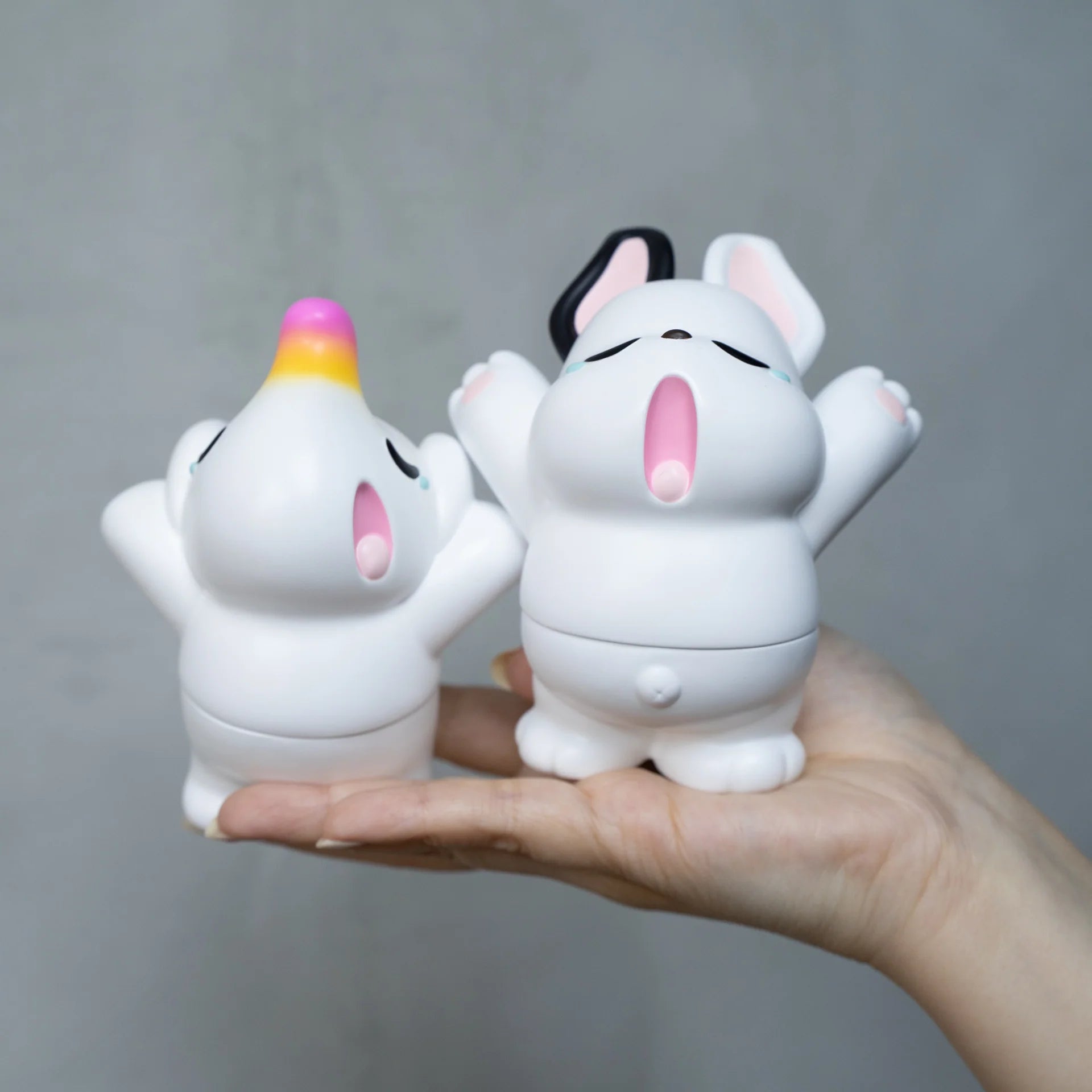 A hand holds two small white animal figurines, Nimbus and Elfie, in Sofubi material. Packaged with a header card, these cute characters yawn, embodying the anticipation of spring.