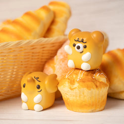 A basket of pastries with a yellow toy elephant, muffins, and a close-up of a cupcake, featuring ELFIE & TEDDY BREAD toy characters.