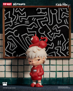 Hirono x Keith Haring Figure: A toy figurine in a red hoodie standing in front of a blackboard. Preorder, ships Aug 2024. Made from PVC/ABS, 10cm tall.