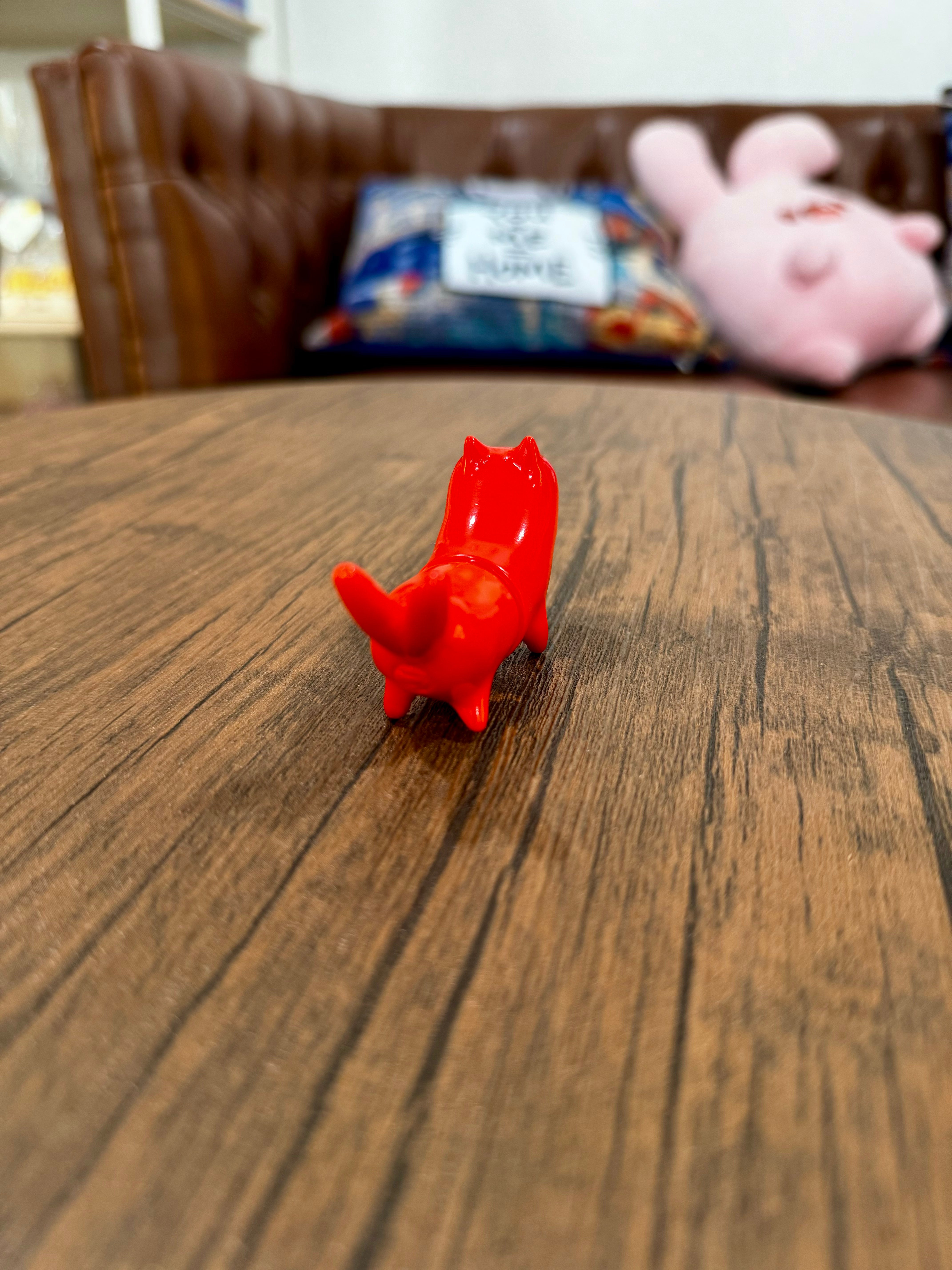 A small red toy cat, Nukomata - Red Sausage by YASU, on a table. Indoor toy with a wooden floor. Reflects Strangecat Toys' blind box and art toy store essence.