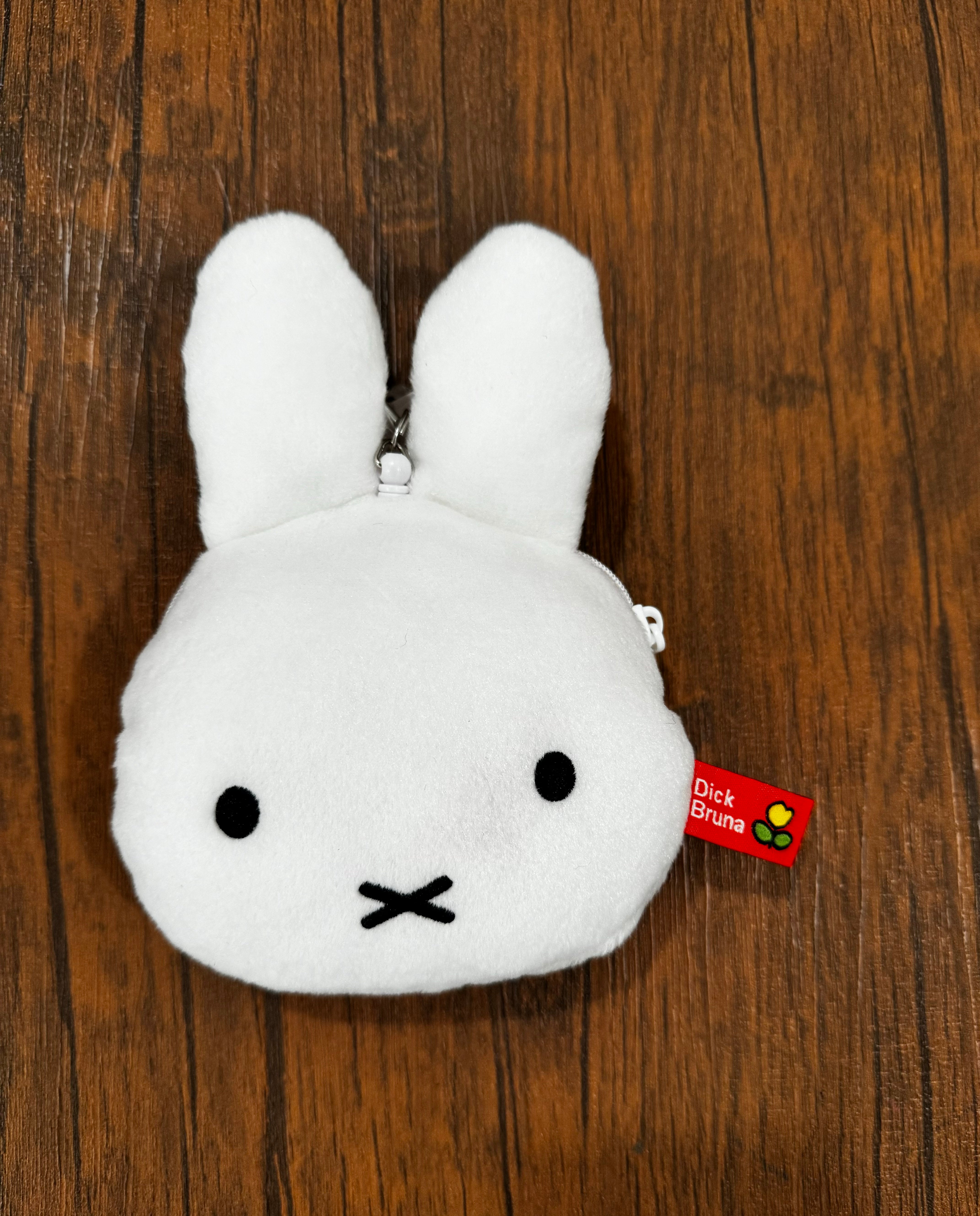 Miffy's face pass case, a white bunny-shaped coin purse, displayed on a wooden surface, featuring a red tag with white text and yellow flowers.