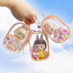 Hand holding a Toy Pochette Keychain with a transparent doll bag and keyring, featuring bells, rings, and hairballs.