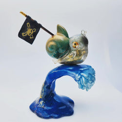 A small figurine of a fish with a flag on top of a wave, a blue and gold slime, a glass figurine of a snail, a black square with a yellow skull and crossbones, and a small pirate flag on a small plastic fish.