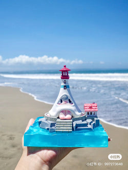 A hand holds Notte Lighthouse by 1001Moons on a beach. Preorder: Ships Late May 2024. Main body 11cm, base 14cm, total height 16cm. Includes lighthouse, island base, lamp set, floor cover.