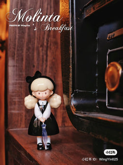 A blind box toy: Molinta breakfast snack figurine, 8cm PVC doll of a girl in a black dress. Cartoonish style, indoor setting.