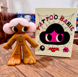 Tattoo Girl - Louise by Burger Babie, a 5 vinyl toy figurine, displayed next to a book featuring a cartoon character. Limited edition of 100 pieces.