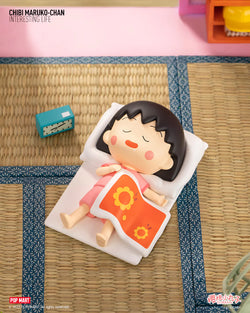 Chibi Maruko-Chan toy doll lying on bed, with a blue clock and yellow ball nearby.