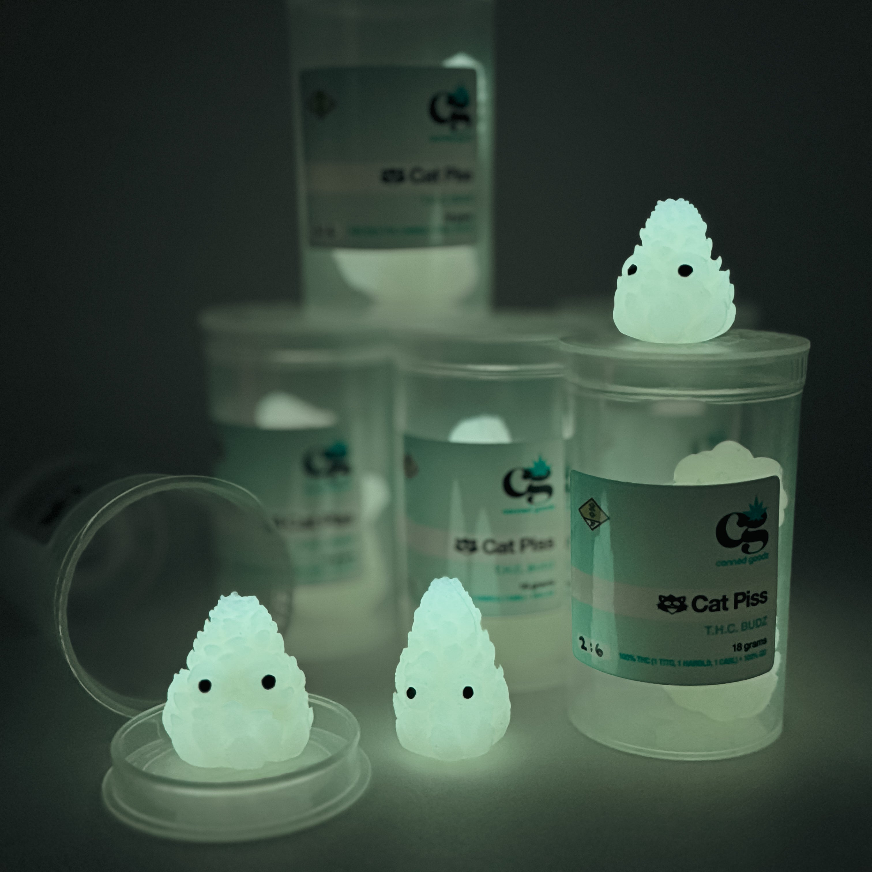 Hello World - THC BUDZ by JFO: A set of three glowing plastic containers, each featuring a white object with black eyes, resembling unique art toys.