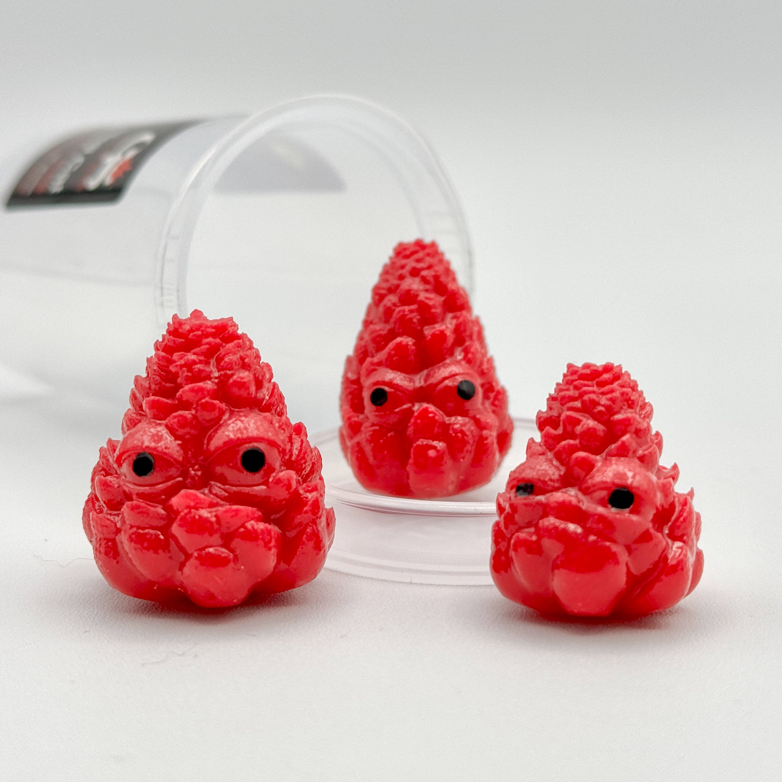 Hello World - THC BUDZ by JFO, a set of three red, toy-like objects with black eyes, resembling berries, displayed for a gallery collection.