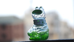 A sofubi/resin BONG A-MON figurine, 9 cm tall, featuring a glass frog design with a green and white scarf. From Strangecat Toys.