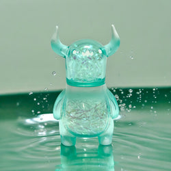 A small plastic Japanese Sofubi toy of Sky Monster Bo by Chishima Konosuke, featuring horns, standing in water, with a penguin-shaped glass container nearby.