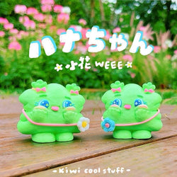 A blind box toy by Kiwi, titled Weee Baby Hey Spring, featuring two green resin toys with pink bows and blue eyes on a wood surface. Size: H5.5cm.