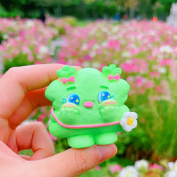 A hand holds a small green toy frog in a field of flowers. Weee Baby Hey Spring by Kiwi, a blind box and art toy store.