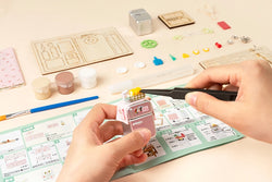 A person holding a toy house kit with intricate details like flowers, spiral stairs, and cozy furniture. From Strangecat Toys, a blind box and art toy store.