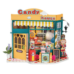 A DIY miniature house kit titled Rainbow Candy House from Strangecat Toys. Imagery includes a toy store setting with candy, a wooden toy clown, and whimsical elements evoking a magical wonderland.