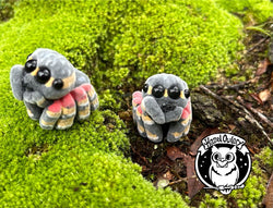 Two stuffed spider toys on mossy surface, one on green plant, and owl logo.