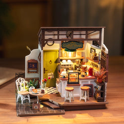 A miniature coffee shop with a table, chairs, a white chair, plant, sign, glass case, and stools from Café Rolife DIY Miniature House Craft Dollhouse.