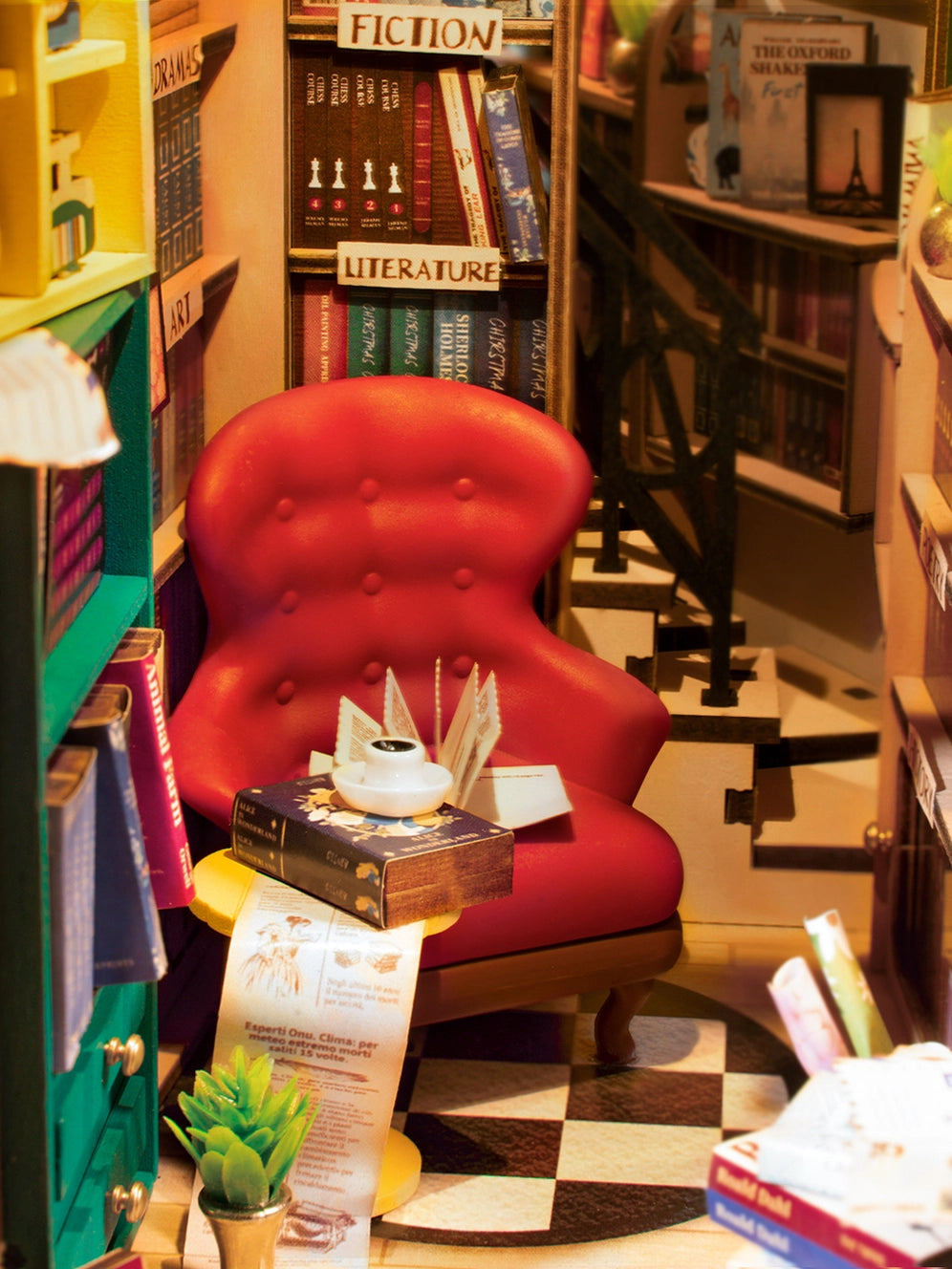 A red chair with books on a table, showcasing Book Nook Kits For Adults - Bookstore. Includes detailed instructions, requires 2 AAA batteries. Dimensions: 9.1 x 12.2 x 1.8 in. From Strangecat Toys.