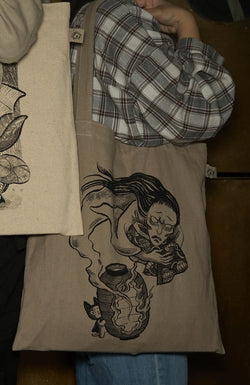 Person holding Badmeaw Yokai Parade Tote Bag with drawing, fabric, and logo details.