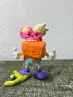 Toy figurine holding cheese, purple object with stem, green and yellow clay figurine, white ball, and more in Strangecat's Halloween Show 2 by Christopher Ian McFarlane.