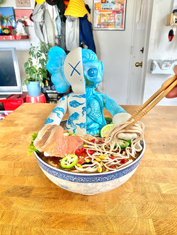 A person holding chopsticks to a bowl of soup with a toy figure, a plant in a blue pot, and a piece of meat on a plate.