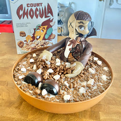 A bowl of cereal with a toy figurine, a chocolate candy, and a chocolate covered dessert.